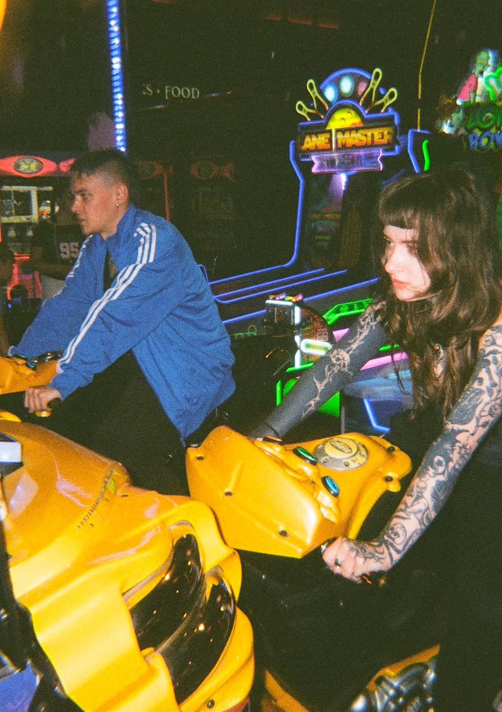 Two people riding yellow arcade motorcycles in a neon-lit arcade, focused expressions, colorful game signs glowing behind them.