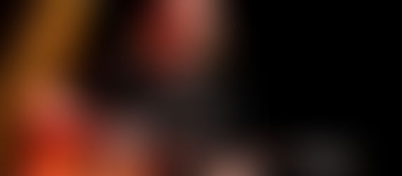 Musician with long hair plays an electric guitar on stage, smiling under a warm spotlight against a dark background.