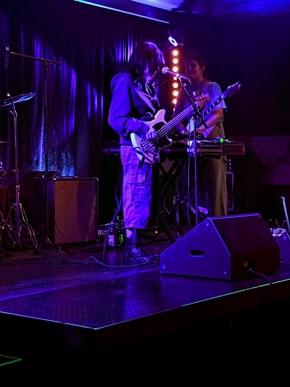 Purple-lit live music stage with two musicians, one playing bass at a microphone and another at a keyboard, cables and amps around them