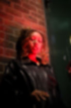 Woman in a black leather jacket leans against a brick wall under red light, eyes closed, with a moody expression.