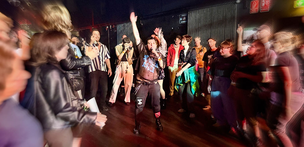 Performer singing to an enthusiastic crowd in a dimly lit venue. People are cheering, with one taking photos. Vivid clothing adds color.