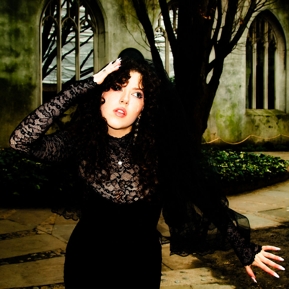 Phoebe Rea in black lace dress poses by a tree and arched windows, gazing off with a moody, dramatic expression.
