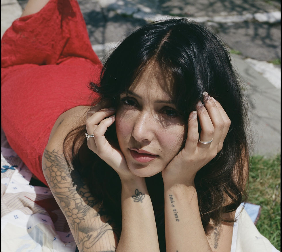 Tattooed woman in a red dress outdoors, hands framing her face, with sunlit grass and the text take time on her wrist.