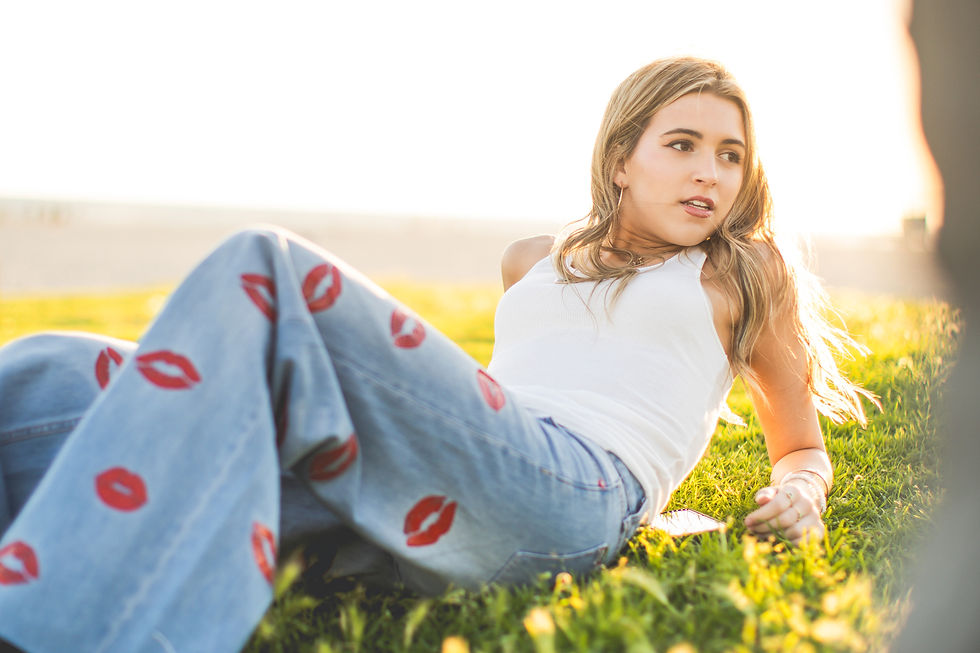 Young woman lounges on sunlit grass, wearing a white tank top and jeans with red lip prints, looking off to the side.