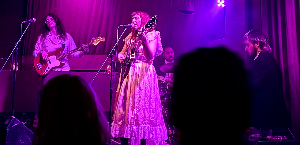 Band performing on stage with purple lighting. Female singer plays guitar, others on bass, drums, and keyboard. Silhouetted audience watches.