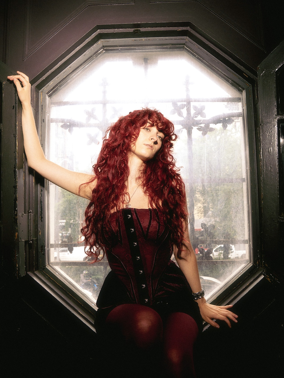 Red-haired woman in a burgundy corset sits by an octagonal window, posing calmly against a bright backlit background.
