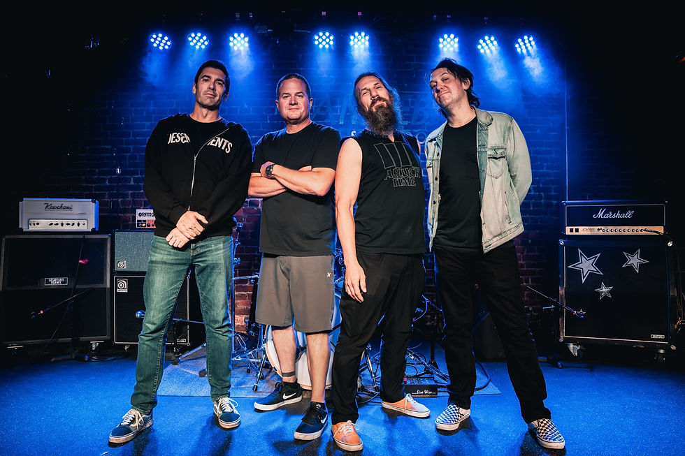 Four men stand confidently on a stage, surrounded by musical equipment. Blue lighting and brick wall background create a vibrant mood.