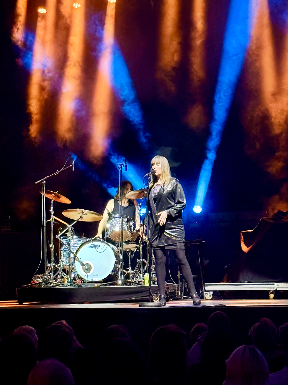 Female singer in black sings into a mic beside a drummer on a concert stage, under blue and orange spotlights.