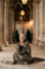 Woman in a grand hallway, wearing a shimmering gold and black gown with intricate patterns, kneels and smiles. Elegant, warm lighting. ESC 2026