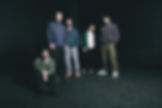 Five men pose in a dark studio, one seated on the floor and four standing in casual clothes against a black background.