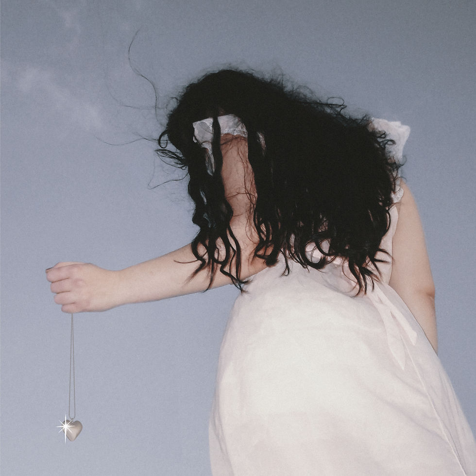 Woman in a white dress with dark windblown hair holds a dangling heart pendant against a plain gray-blue background.