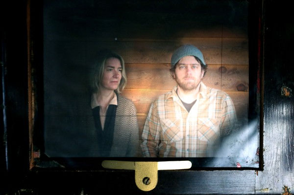 A woman and man sit in a dimly lit room with wooden walls. The man wears a gray beanie and plaid shirt. Both appear contemplative.