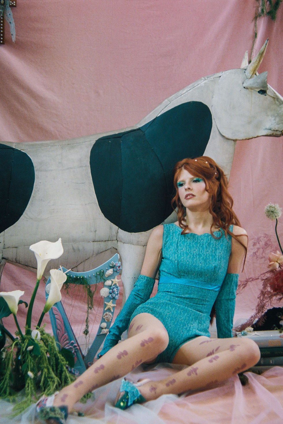 Red-haired woman in a turquoise dress sits before a giant unicorn prop and pink backdrop, posing calmly in a dreamy setting
