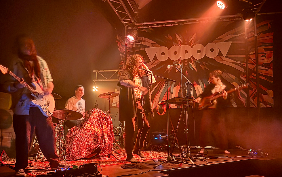 Rock band performs under orange lights on a stage with a Voodoo banner; singer at mic, guitarists and drummer playing.