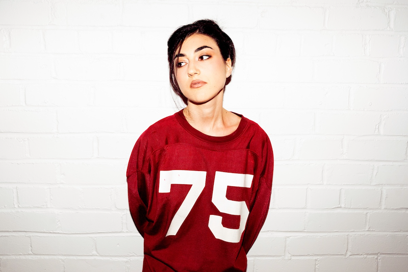 Young woman in a red 75 sweatshirt stands against a white brick wall, looking off to the side with a pensive expression.