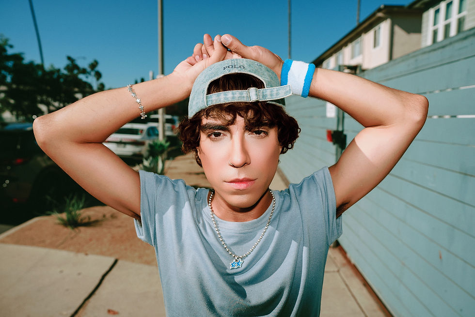 Young man in a blue shirt and cap poses with arms raised on a sunny street, wearing jewelry and a polo logo cap.
