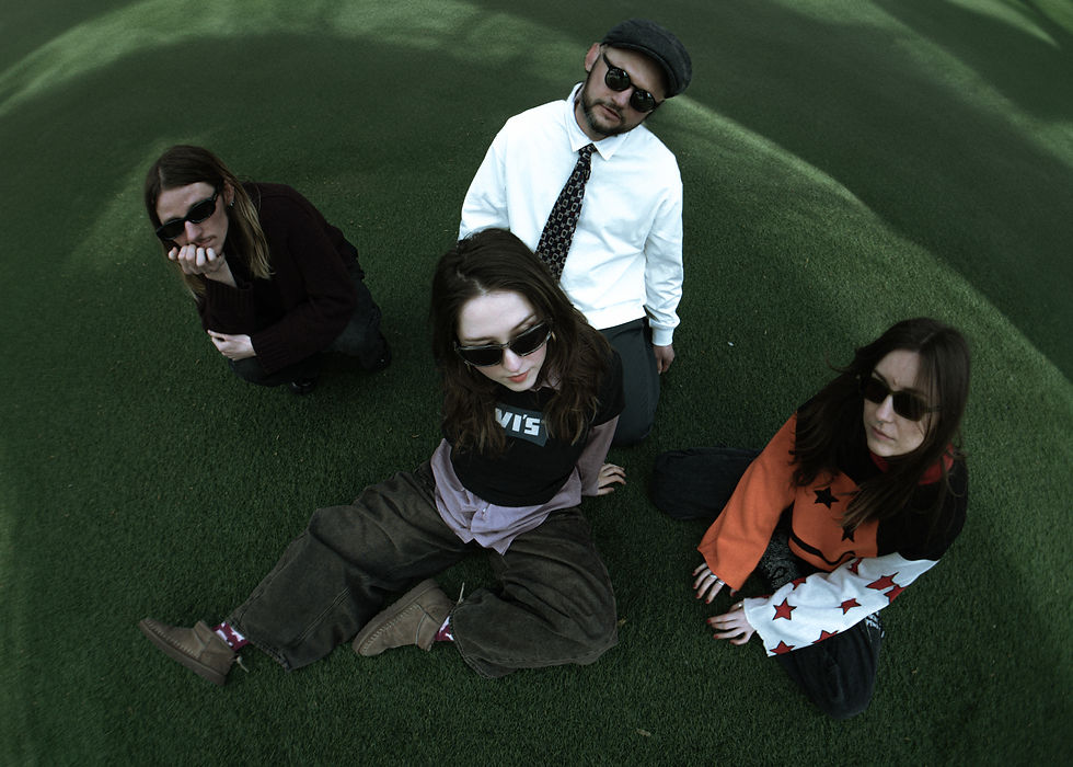 Four people wearing sunglasses sit on grass, looking up. One wears a white shirt and tie, another a Levi's shirt. The mood is relaxed.