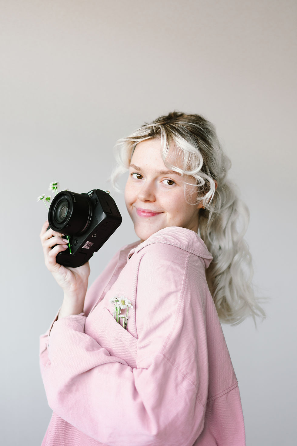 Isobel Buxton Photography smiling in front of blank white wall wearing pink shirt and camera adorned with flowers.
