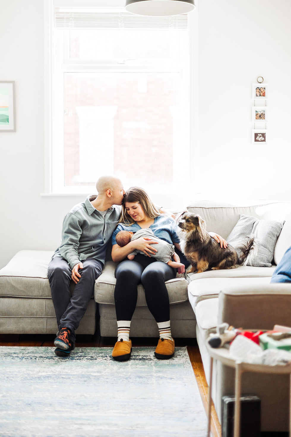 A couple sits on a sofa with a baby and dog. The man kisses the woman's head. Bright room, cozy vibe, natural light through a window.