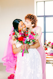Two brides smiling and hugging with a bouquet bursting with pink, red and greens at Brickhouse Vineyard, Exeter, Devon, UK Wedding venue