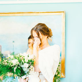 Bridesmaid crying in front on light blue wall while holding a bouquet of lush foliage and white florals at Deer Park wedding venue, Devon.