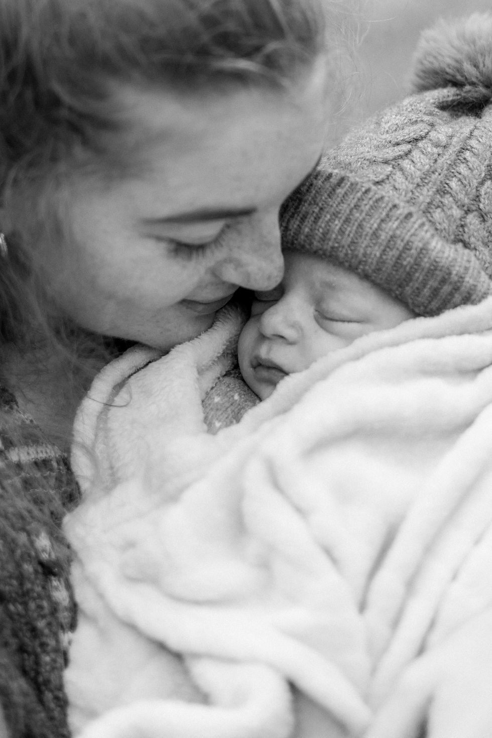 Tender black-and-white image of a woman gently nuzzling a sleeping baby wrapped in a soft blanket, wearing a knit hat. Peaceful and intimate mood.