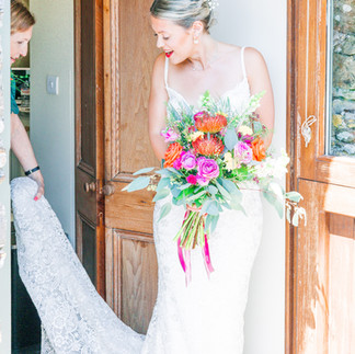 Bride walking through door frame while holding bright bouquet. Her mother is holding her dress train behind her