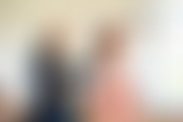 Woman with orange curly hair wearing a pink suit is having hair spray in her hair by a hair stylist