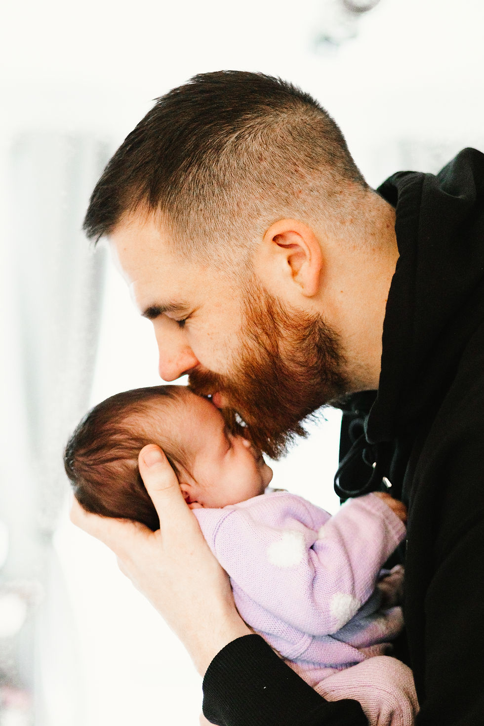 Man gently holds and kisses newborn's forehead, creating a tender moment. Baby in purple outfit. Bright, soft background conveys warmth.