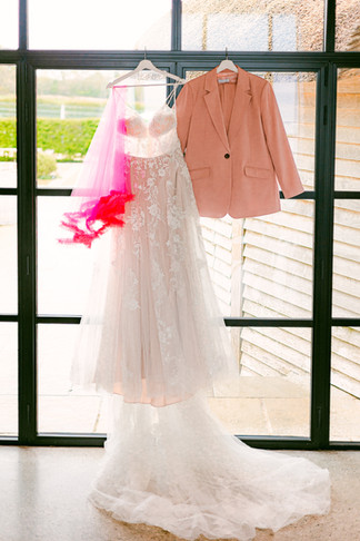 Wedding dress hung up on a large window next to a hot pink veil and a pink suit at Brickhouse Vineyard, Exeter, Devon