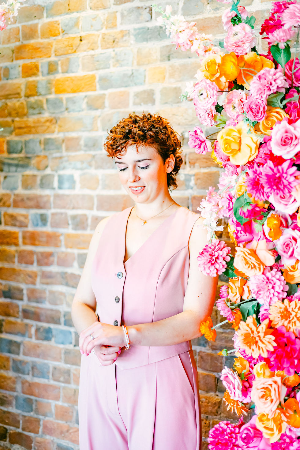 Woman in a pink outfit stands beside a vibrant floral arrangement against a brick wall, looking down with a serene expression.