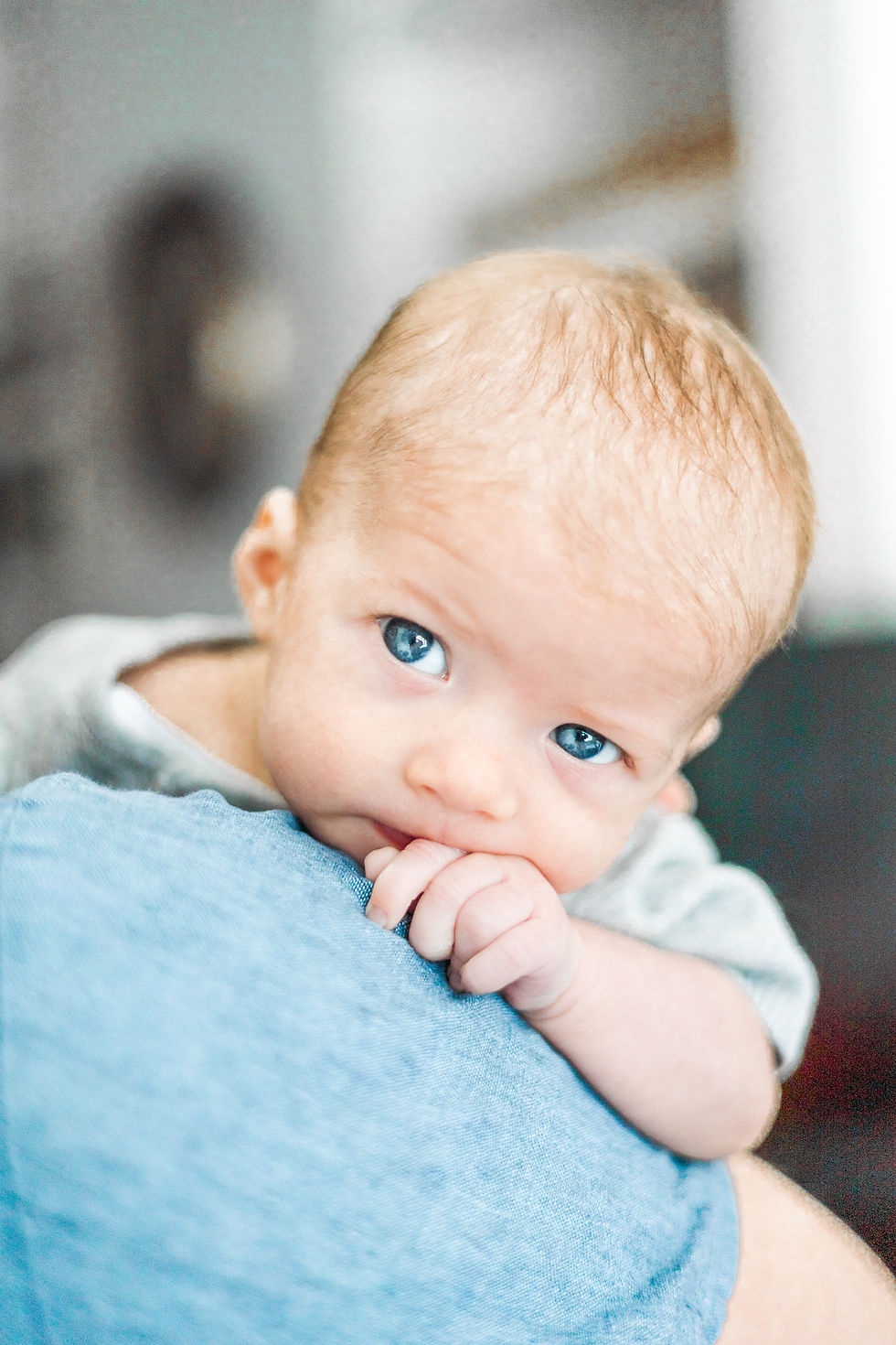Baby with blue eyes rests on a blue-clad shoulder, gently chewing fingers. Soft lighting creates a warm, serene atmosphere.