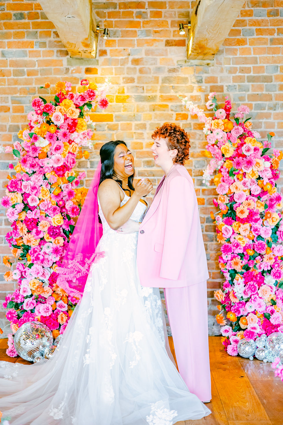 Two joyful women, one in a dress with a pink veil, the other in a pink suit, laughing against a brick wall backdrop adorned with vibrant flowers at Brickhouse Vineyard, Exeter, Devon, UK wedding venue