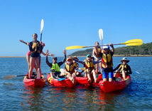 A group of kayaks, kids standing or kneeling, raising paddles, everyone laughing