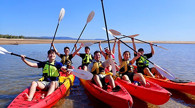 A group in life jackets raising their paddles from red kayaks gathered on calm water.