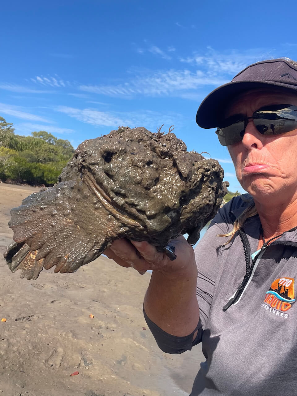 Person holding a large, muddy stonefish, making a pouting face. Background shows a sunny blue sky and a sandy area with trees.