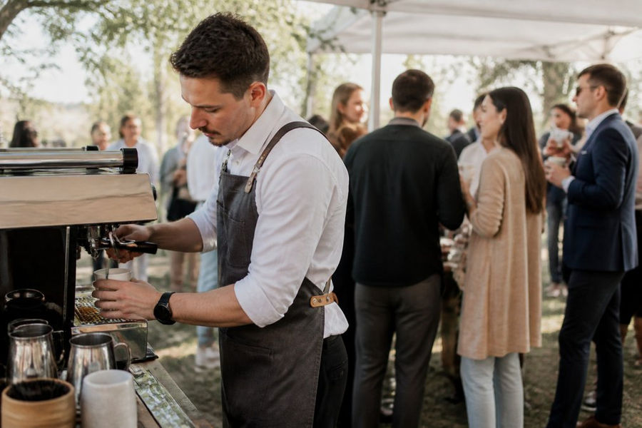 Professional barista preparing handcrafted espresso drinks at an outdoor event with guests enjoying beverages in natural afternoon light