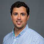 A man in a blue and white checkered shirt smiles for a corporate team headshot in the Carlsbad office setting.