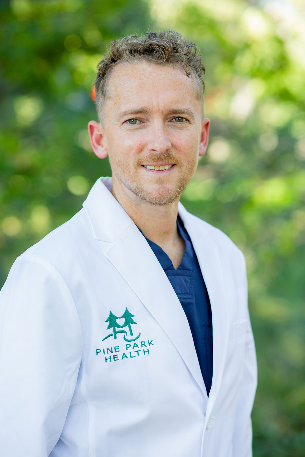 Smiling male physician assistant -certified in a white lab coat with "Pine Park Health" logo, outdoors with green blurred background in San Diego for headshots.