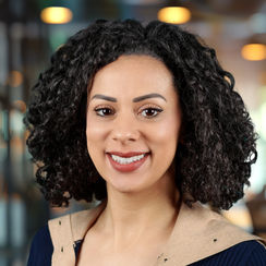 A woman with curly hair in a blue shirt poses for a professional headshot in an office environment.