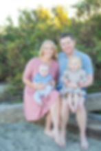 A smiling family of four sits on a wooden log at the beach. Parents wearing pink and blue hold young children. Lush greenery in background.