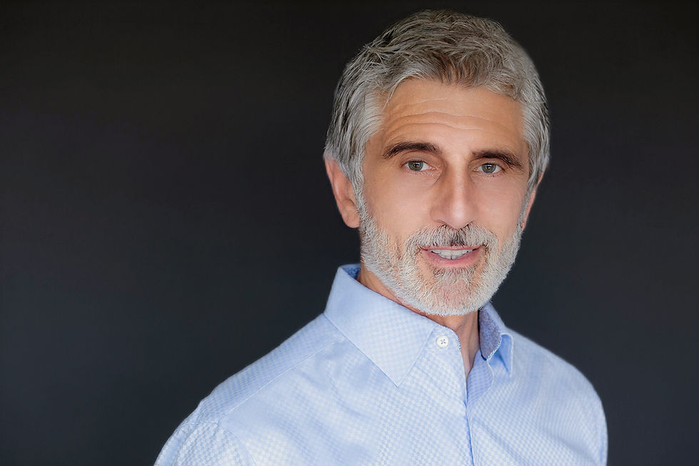 Man in his 60's wearing a light blue dress shirt with a dark grey background, smiling for a headshot photo