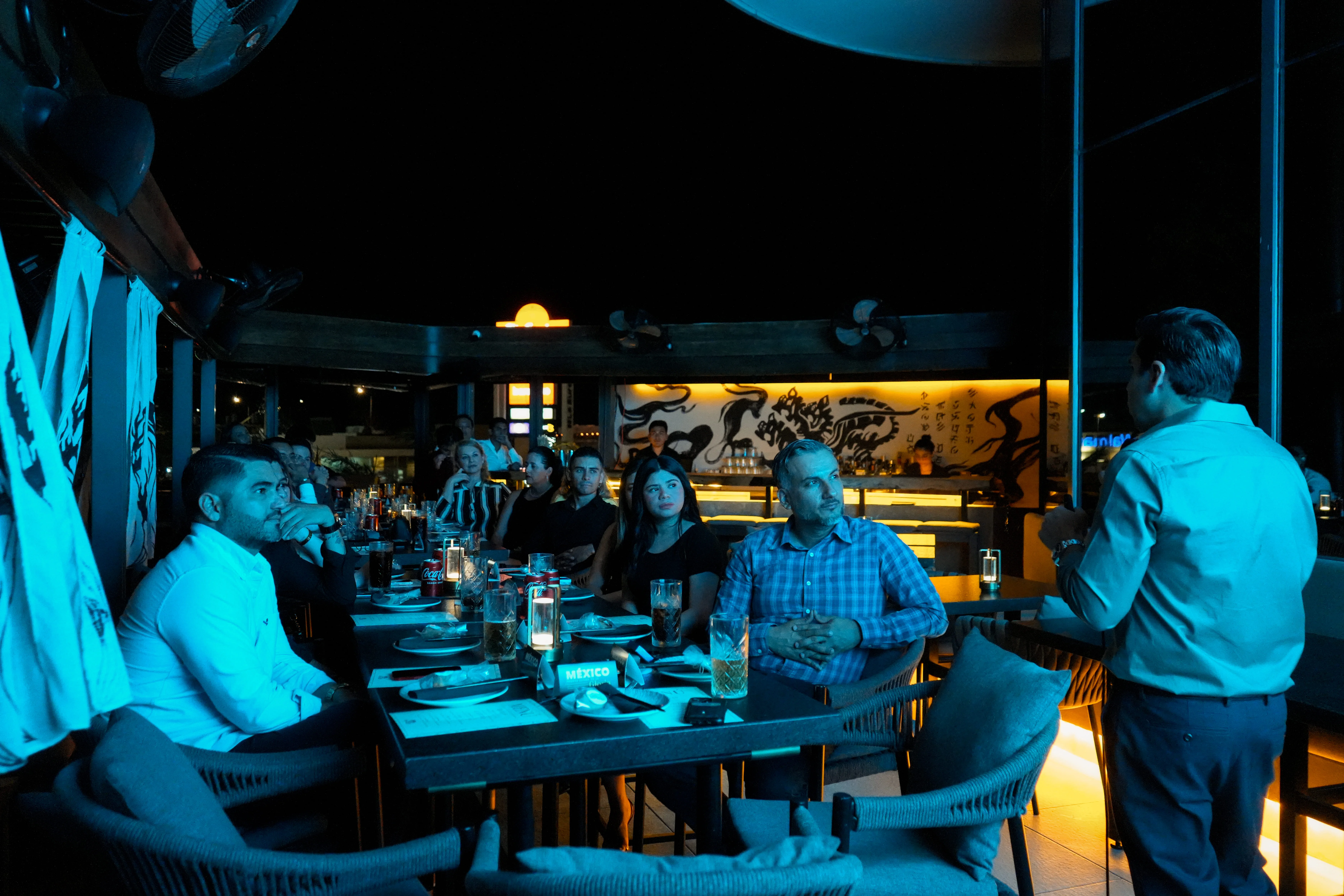 sujeto hablando hacia mesa con multitud de personas de noche luz azul