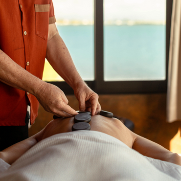 Spa therapist applying warm stones to person's back, ocean view.