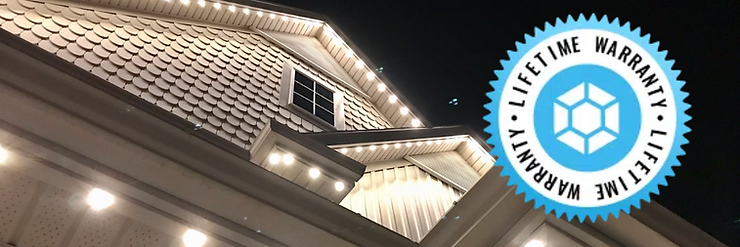 Nighttime view of a well-lit house roof with decorative shingles, and a "Lifetime Warranty" badge in blue and white on the side.