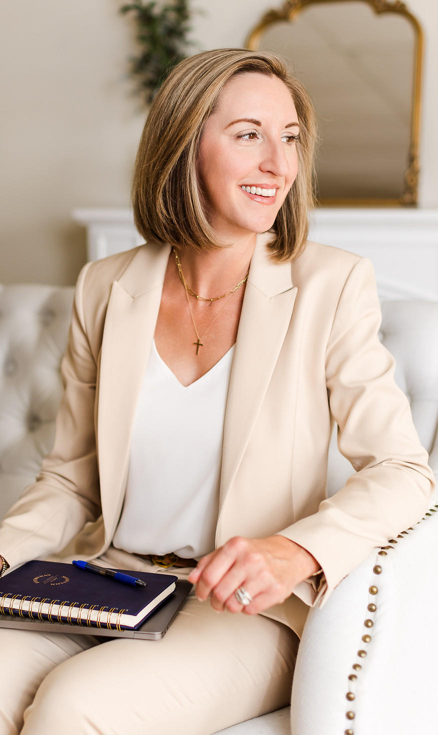 A smiling Lesley Herald, a blonde woman, looks off to the right. She is wearing a tan pantsuit and a white top, representing her "people first" core value. She holds a dark blue notebook and a pen, with a white tufted couch and a gold mirror visible in the background.