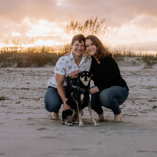 Family of two humans and one dog huddle together smiling and posing for a beach family photo session with Wilmington's pet photographer, The Photo District.