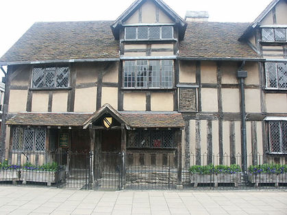 Zdjęcie przedstawia zabytkowy, drewniano-ceglany dom w stylu Tudorów, znany jako miejsce narodzin William Shakespeare. Budynek ma charakterystyczną konstrukcję szachulcową z ciemnymi belkami i jasnym wypełnieniem, dwuspadowy dach oraz niewielkie okna z podziałami. Przed domem znajduje się niewielki ogród oraz ogrodzenie. Całość ukazuje historyczną zabudowę miasta Stratford-upon-Avon i ma charakter muzealny.