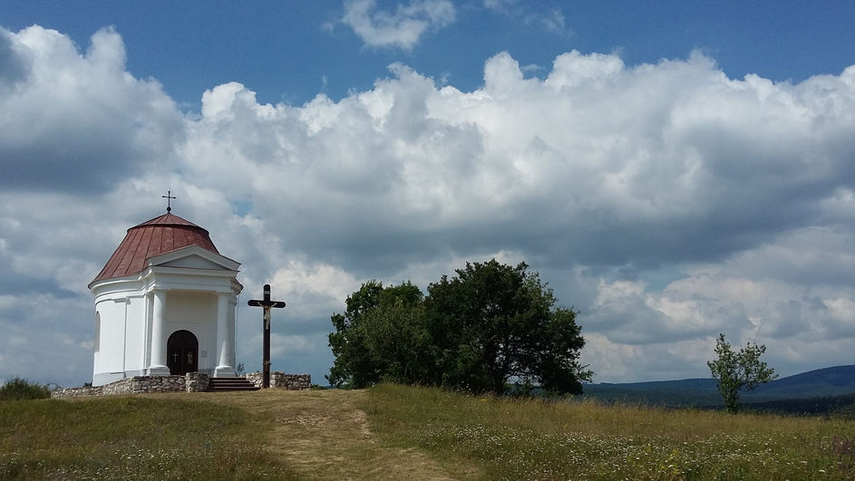 Fehér kápolna és fa kereszt dombtetőn, felhős ég alatt, mezőn.