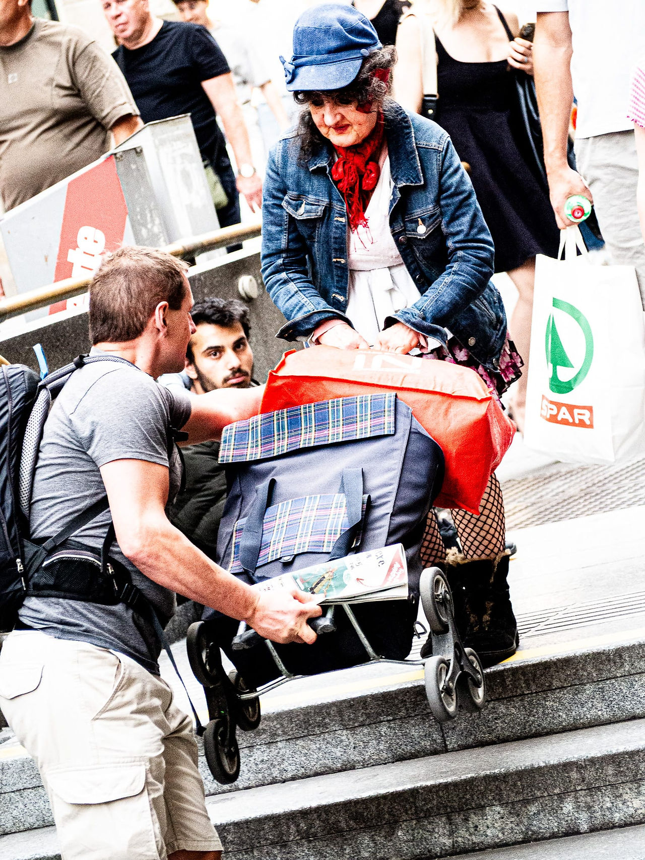 Woman helping man with bags on stairs, people in background, shopping bag.